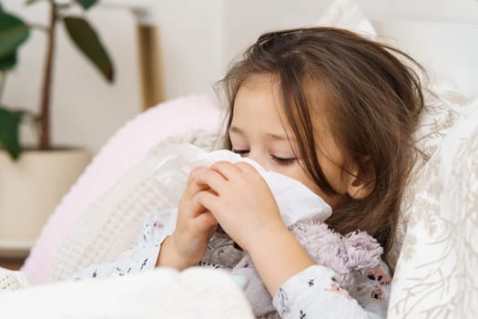 Sad, depressed little long haired girl covering face with napkin tissues, lying with soft toy, wrapped in blanket. Flu season, coronavirus and allergy sypthoms with seasonal infection. Bedtime at home