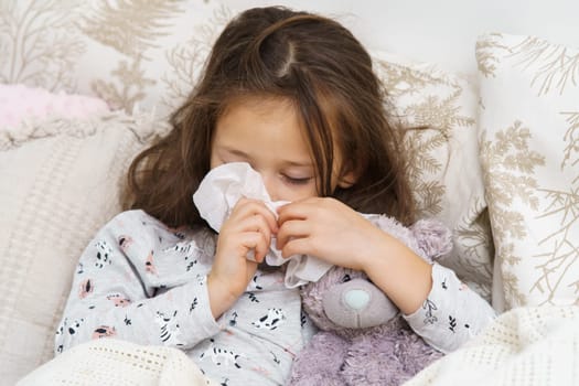 Tired, sleepy little brunette girl sneezing snot with tissues, embracing bear toy in soft bed. Headache and rhinitis symptoms with seasonal viral infection. Bedtime at home, lockdown and quarantine
