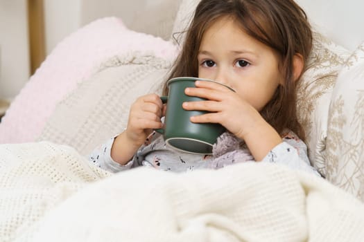Nice, sweet, dear little dark haired preschooler girl drink hot tea or medicine from green mug, lying in pajamas in soft bed. Recovering, health care. Taking medicine, drinking dissolved medicine