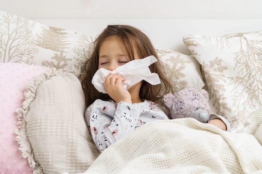Sleepy, exhausted little brunette girl with close eyes wiping wet nose with tissues, lying with bear toy in bed. Flu season and rhinitis, headache and allergy with seasonal infection. Fever and cold