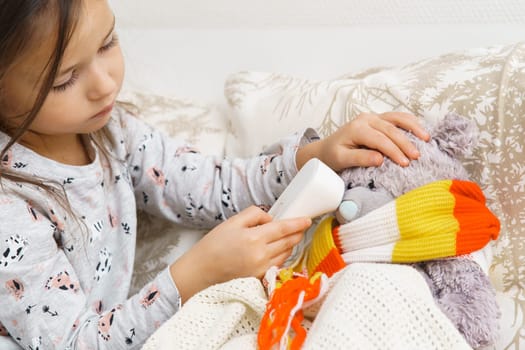 Concentrated child, girl healing bear toy, examining temperature with electronic thermometer, wrapping in scarf on bed, sit on sofa. Illness, sickness and seasonal flue. Little doctor help best friend