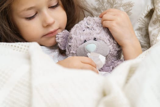 Cute little brunette girl healing, wiping imaginary snot of bear toy patient with tissues, lying, wrapped in blanket. Flu season ahead, headache and allergy with seasonal infection. Bedtime at home