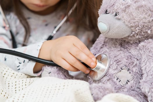 Unrecognizable small girl listen to bear toy patient heartbeat with stethoscope, wrapped in blanket. Little doctor help best friend. Loneliness and fear during disease at home. Healthcare and medicine