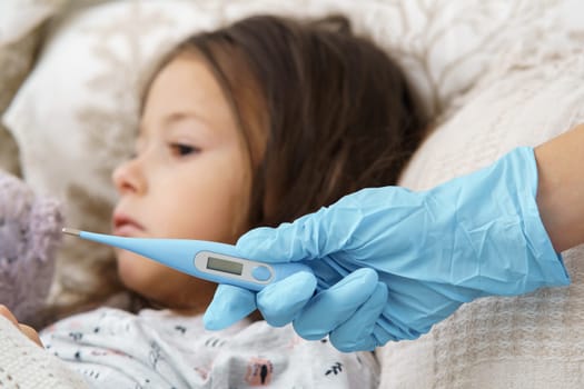 Cropped doctor hand in glove holding electronic thermometer against calm, sad, silent little girl. lying in bed. Unwell condition and viral infection. Quarantine, lockdown during covid 19. Bed rest