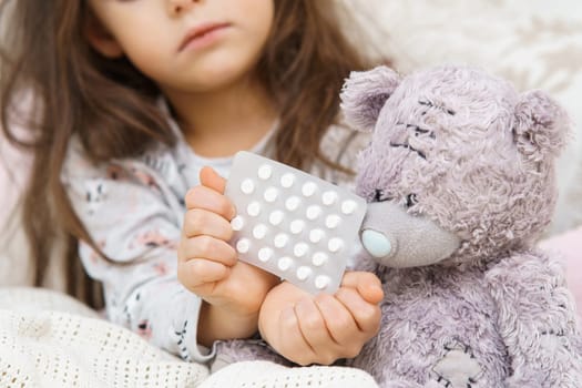 Cropped little brunette girl hands holding pills blister near bear toy patient with sit on bed in bedroom. Loneliness and fear during disease. Child sickness, unwell condition. Pharmaceutical recovery