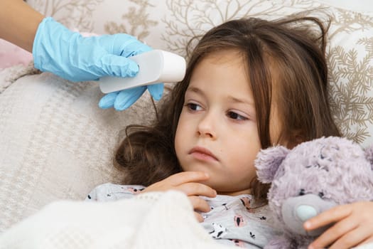 Cropped doctor hand in glove using electronic thermometer against feared, sad, silent brunette girl. lying with toy in bed. Unwell condition, viral infection. Bed rest, illness and inflammation