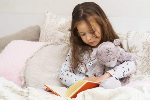 Little pensive, nice lonely brunette girl in pajamas reading book with teddy bear on soft bed. Imagination and day dreaming. Learning at home, education and knowledge. Leisure activity during lockdown