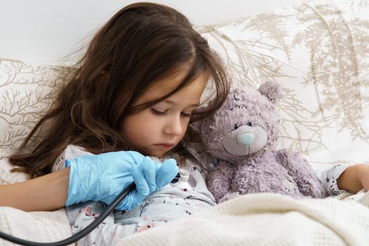 Cropped doctor hand in sterile glove using stethoscope to listen to heart and lungs of little girl lying in bed with bear toy. Seasonal infection, virus, coronavirus. Healthcare and medicine