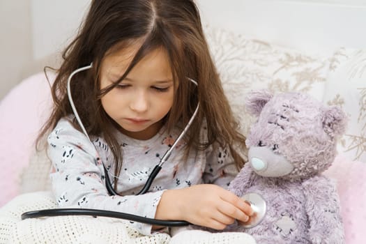 Serious, concentrated, careful small child, girl checking teddy bear chest, breathing with stethoscope, wrapped in blanket. Healthcare and medicine. Little doctor help at home. Friends sickness