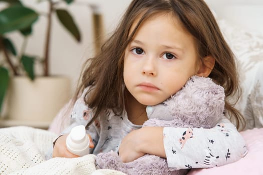 Sad depressed worried, sunless little brunette girl embracing bear toy, healing with nose spray, sit on bed and look at camera. Kid sickness, virus and covid 19. Lockdown isolation, loneliness at home
