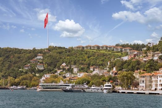 View from the sea of the green mountains of the Europian side of Bosphorus strait, with docked boats, traditional houses, dense trees, and huge Turkish flag, in a summer day