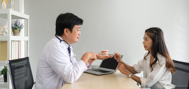 Asian dentist in white gown and stethoscope pointing finger at tooth model, explain to young patient about orthodontics and braces. Help reduce gum pain. Healthcare and medicine concept.