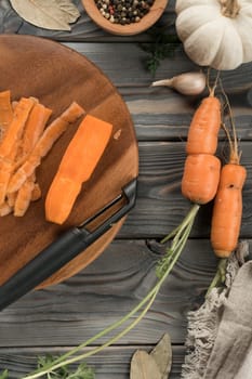Peeling ripe orange carrot with vegetable peeler. Flat lay view. Fresh peeled carrot. Cutting peel on kitchen wooden board. Prepare ingredients for cooking, food waste. Vegetables and flavorings.