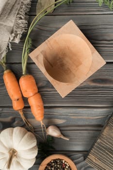 Ripe unpeeled orange carrot with green haulm. Empty bowl on kitchen wooden table. Flat lay view. Layout of fresh vegetables and flavorings. Prepare ingredients for cooking. Healthy food.