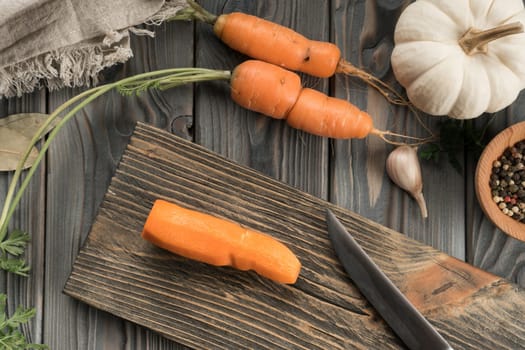 Cut ripe raw orange carrot on kitchen wooden board with knife. Whole peeled carrot. Flat lay view. Layout of fresh vegetables and flavorings. Prepare ingredients for cooking. Healthy food.
