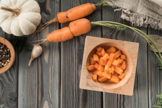Cut ripe raw orange carrot into cubes. Diced carrot in bowl on kitchen wooden table. Flat lay view. Layout of fresh vegetables and flavorings. Prepare ingredients for cooking. Healthy food.