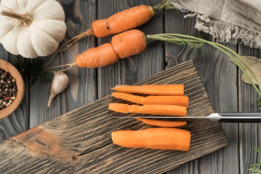 Cut ripe raw orange carrot into slices on kitchen wooden board. Sliced carrot with knife. Flat lay view. Layout of fresh vegetables and flavorings. Prepare ingredients for cooking. Healthy food.
