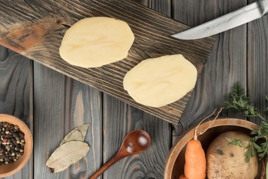 Cutting in half raw yellow potato with knife on kitchen wooden board. Fresh sliced potato, traditional cuisine. Flat lay view. Ripe vegetables and flavorings. Prepare ingredients for cooking.