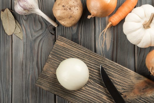 Cutting spicy onion with knife on kitchen wooden board. Whole white onion. Flat lay view. Layout of fresh raw vegetables on table. Prepare ingredients for cooking. Healthy diet, foodstuffs.