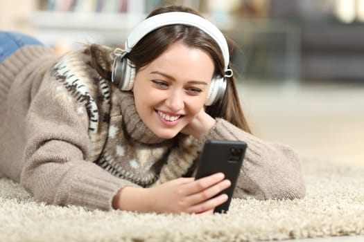 Happy teen in winter watching media on phone wearing headphones on the floor at home