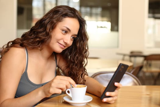 Woman checking smart phone and stirring coffee sitting in a bar