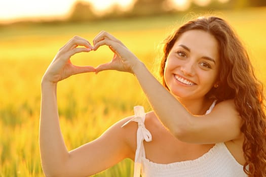 Happy woman making heart shape with hands at sunset in a wheat field