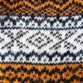 A fragment of an ornament on a knitted woolen sweater. Winter motives. Beige, brown, white, black threads for hand knitting. Knitted pattern. Rhombuses, snowflakes, lines, waves, dots and circles.
