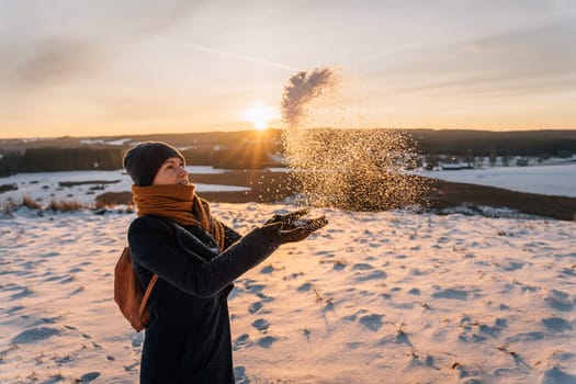 A woman in a snow-covered field tosses snow with her hands against the backdrop of a sunset.