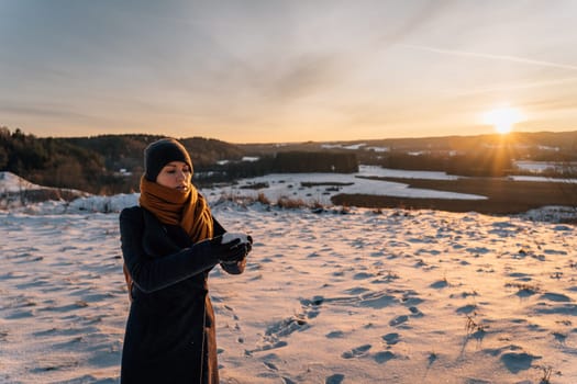 A young woman in a coat and hat at sunset stands on a snow-covered field and holds a handful of snow in her hands.