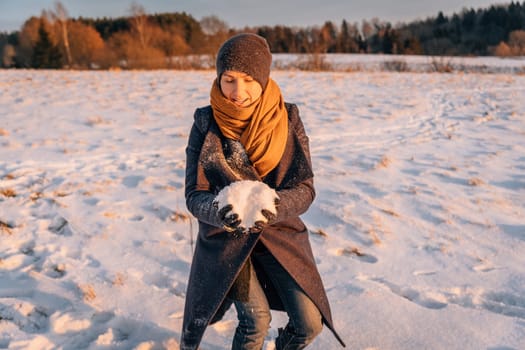 A woman in winter clothes collects a lump of snow in a snow-covered field.