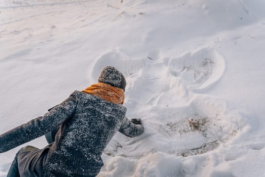 A woman in a snow-covered coat and hat draws a snow angel in the snow.