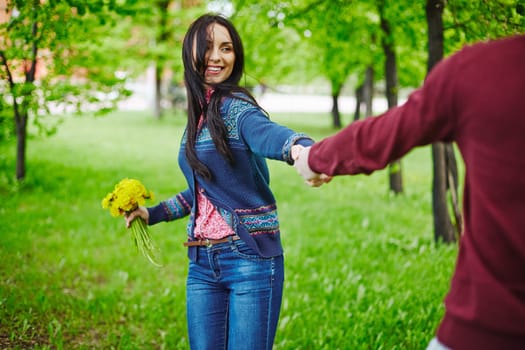 Smiling girl with bunch of yellow dandelions holding her boyfriend by hand in park