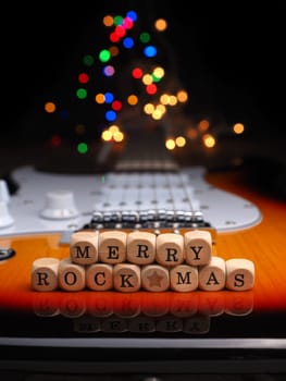 Wooden blocks with the inscription Merry Rock Mas on an old used vintage guitar with colorful Christmas lights bokeh background