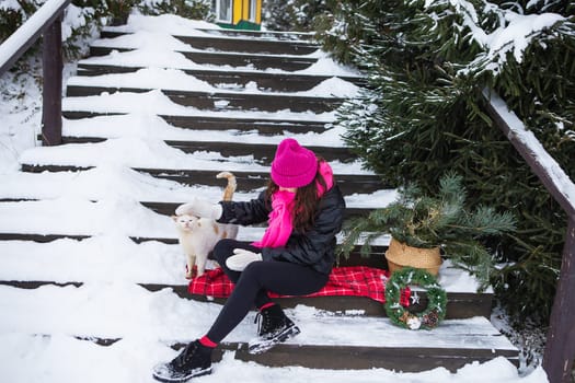 Woman holds and strokes white furry cat near Christmas tree. Domestic pet enjoys affection and attention. New Year's magical atmosphere of comfort, love and warmth.