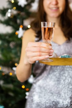 A girl in a shiny dress holds a glass of champagne against the background of a decorated Christmas tree, preparing for the new year and christmas