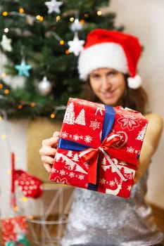A girl in a shiny dress and a santa claus hat holds a beautifully sealed gift against the background of a decorated christmas tree, preparing for the new year and christmas