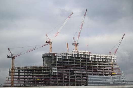 Workers are busy on a construction site in Paris, where a high-rise building is taking shape under a cloudy sky with cranes lifting materials.