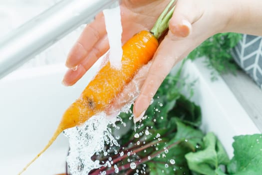 Cropped photo of woman washing orange carrot with leaves under running splashing water from brass faucet in kitchen. Vegetables, vegetarian, preparation for cooking, healthy food, hygiene, vitamins.