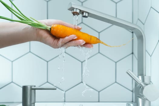 Side view of unrecognizable woman washing orange carrot with leaves under running water from modern brass faucet in kitchen. Vegetables, vegetarian, preparation for cooking, healthy food, hygiene.
