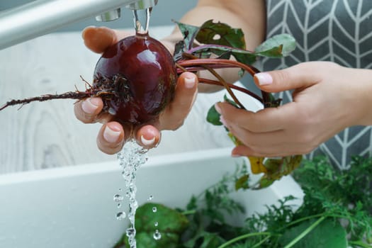 Cropped photo of woman wearing grey apron washing fresh beet with hands under running water in sink full of greenery in kitchen. Vegetables, vegetarian, preparation for cooking, healthy food, hygiene.