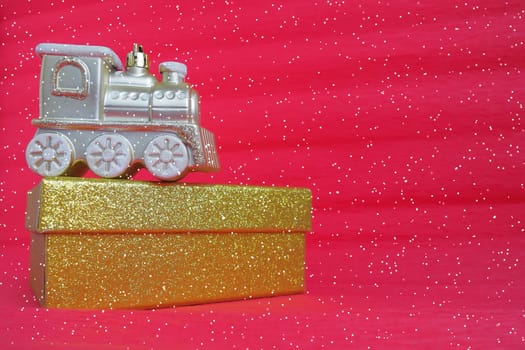 Golden toy train locomotive and gift box on a red gradient background. New Year or Christmas card. Icicles and snow on a train. Free space for text. Copy space. Falling snowflakes