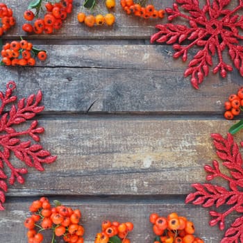 Red, orange and yellow winter rowan berries on a horizontal wooden background. Red shiny large snowflakes are arranged in circle around the edges background. New Year or Christmas natural backdrop.