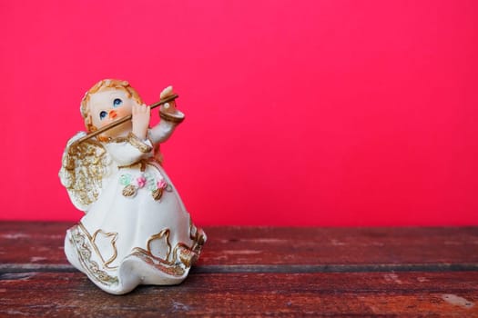 Guardian angel plays the flute. Figurine of a red-haired girl in a white dress with wings and a pipe. New Year or Christmas holiday background. Copy space. Pink wooden background. Free space for text