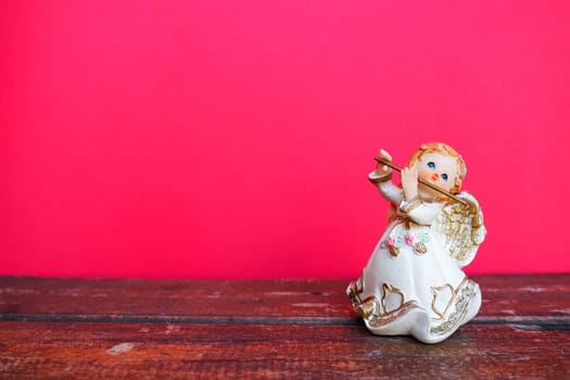 Guardian angel plays the flute. Figurine of a red-haired girl in a white dress with wings and a pipe. New Year or Christmas holiday background. Copy space. Pink wooden background. Free space for text