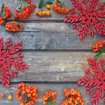 Red, orange and yellow winter rowan berries on a horizontal wooden background. Red shiny large snowflakes are arranged in circle around the edges background. New Year or Christmas natural backdrop.