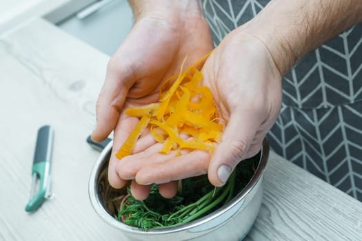 Cropped photo of man wearing grey apron holding carrot peelings in hands over metal bowl full of peelings near peeler, standing at grey wooden table in kitchen. Recycling, composting, fertilizing.