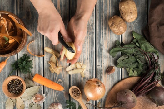 Unrecognizable woman peeling potato with black knife near brown wooden bowl with carrot peelings, little bowl with peppercorns, peeled carrot, onion, garlic, beets, bay leaves on grey wooden table.