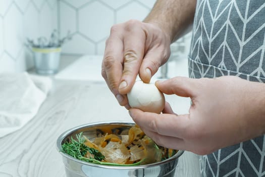 Cropped photo of man wearing grey apron peeling hardboiled egg over metal bowl full of carrot, onion peelings, standing at grey wooden table in kitchen. Recycling, composting, fertilizing, zero waste.