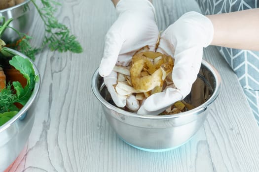 Cropped photo of man wearing grey apron, white disposable gloves, holding potato peelings in hands over metal bowl full of peelings, standing at grey wooden table in kitchen. Recycling, composting.