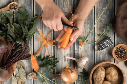 Unrecognizable woman peeling carrot with black knife near brown wooden bowl with potatoes, little bowl with peppercorns, tiny grater, onion, garlic, beets, bay leaves on grey wooden table. Top view.